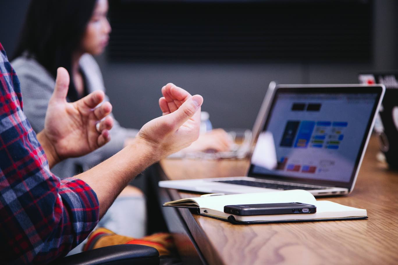 Shows a meeting, focused in on one persons hands and laptop, with others in the background